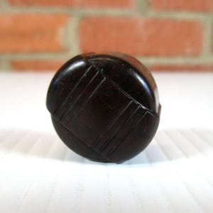 Op de afbeelding: Een donkerbruine, ronde knoop met een gesneden, geometrisch ontwerp. De knoop staat op een wit oppervlak, met een vage bakstenen muur op de achtergrond. Het oppervlak van de knoop is glad en reflecterend.