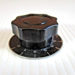 Może przedstawiać: Czarny, ośmiokątny guzik z okrągłą podstawą z białymi cyframi i oznaczeniami. Pokrętło służy prawdopodobnie do regulacji ustawień w urządzeniu. Podstawa ma cyfry od 0 do 5.