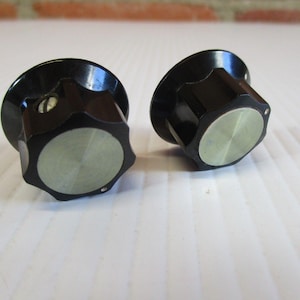 May include: Two black control knobs with a silver-colored center. The knobs have a faceted edge and a circular, brushed metal center. They are likely for use on electronic equipment.
