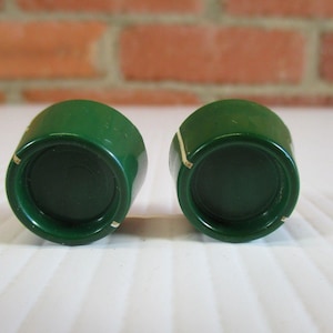 May include: Two vintage, dark green knobs with a rounded top and a flat, circular base. The knobs have a glossy finish and are set against a white surface with a brick background. Each knob has a small, light-colored mark.