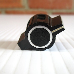 May include: A black control knob with a textured circular center and a silver metallic ring border. The knob features a protruding wing-shaped handle on one side with a thin silver accent line running along the top edge of the wing extension.