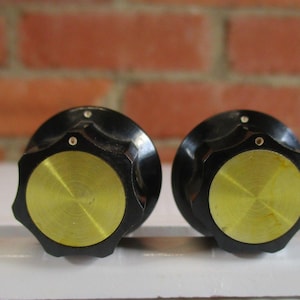 May include: Two circular control knobs featuring a black fluted outer edge and a flat, circular brass-colored center inlay. Each knob has a small white dot indicator on the top edge of the black rim, providing a clear visual reference point.