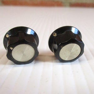 May include: Two black, vintage-style knobs with a silver center and a star-shaped edge. These knobs are likely for a stereo or other electronic device. The knobs are set against a white background.