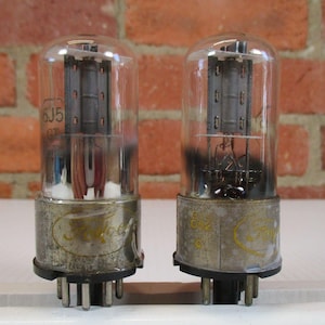 May include: Two vintage vacuum tubes with glass enclosures and metal bases. The tubes have internal components visible through the glass. The bases are silver with gold lettering. The background is a brick wall.