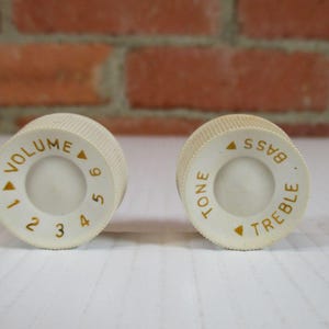 May include: Two off-white guitar knobs with gold lettering. One knob reads "VOLUME" with numbers 1-9. The other reads "TONE", "BASS", and "TREBLE". The knobs are set against a brick wall.