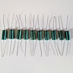 Puede incluir: Una colección de diez condensadores electrónicos cilíndricos de color verde azulado dispuestos en una fila horizontal. Cada componente presenta un cuerpo metálico con texto impreso y dos cables conductores finos y rectos.
