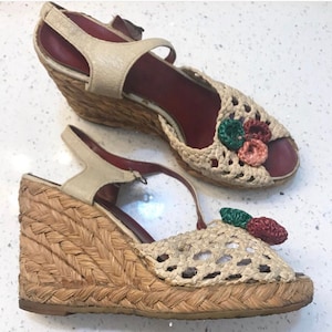 Op de afbeelding: Een paar geweven espadrille-wigsandalen met een gehaakt bovenwerk en bloemversieringen. De sandalen zijn beige met een bruine wigzool.