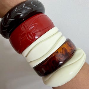 May include: A collection of stacked bangles in various colors and designs. The bangles include a dark brown carved leaf design, a red carved flower design, white wavy bangles, a brown tortoiseshell bangle, and a cream-colored wavy bangle.
