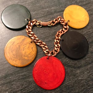 May include: A necklace featuring a copper chain with five large, round pendants in various colors: green, yellow, black, red, and a marbled orange. The pendants are connected to the chain by small loops.