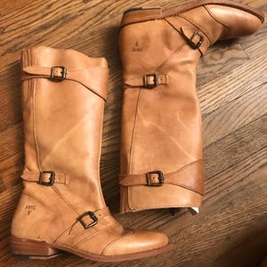 Könnte beinhalten: Ein Paar kniehohe Lederstiefel der Marke Frye in Hellbraun. Die Stiefel haben mehrere verstellbare Riemen mit Metallschnallen und einen niedrigen Absatz. Das Frye-Logo ist auf der Seite jedes Stiefels sichtbar.