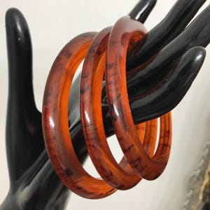 Peut inclure: Quatre bracelets translucides orange et marron. Les bracelets ont un aspect marbré et sont présentés sur un présentoir noir en forme de main. Les bracelets sont de couleur chaude et ont une finition brillante.