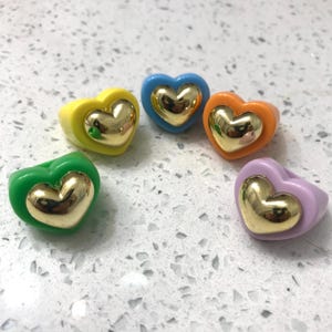 Puede incluir: Cinco anillos en forma de corazón con detalles dorados. Los anillos son de diferentes colores: verde, amarillo, azul, naranja y morado.