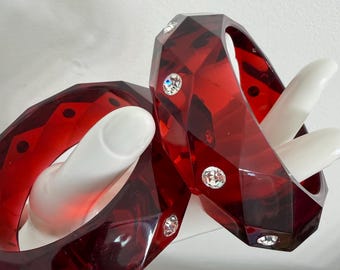 May include: Two faceted, translucent red bangles with embedded clear crystals. The bangles are held by a white hand. The bangles have a geometric design and are a deep red color.