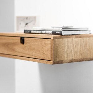 May include: A light wood floating shelf with a drawer and a stack of books on top. The books are titled "Michelangelo Antonioni" and "Taschen".