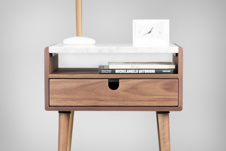 May include: A wooden nightstand with a white marble top, a drawer, and a shelf. The nightstand has four legs and a clock on the top shelf. The books on the shelf are titled "Dior Michelangelo Antonioni" and "Taschen".