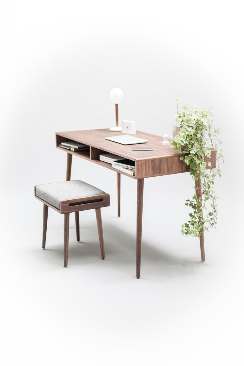 May include: A wooden desk with a light wood finish and a gray upholstered stool. The desk has a shelf and a drawer. A lamp, a laptop, and a plant are on the desk.