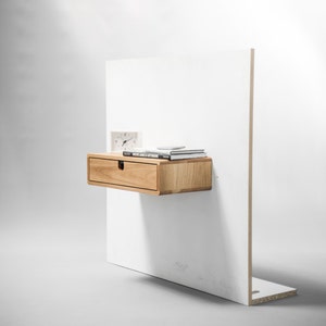 May include: A light wood floating nightstand with a single drawer and a white wall behind it. The nightstand has a clock and books on top.