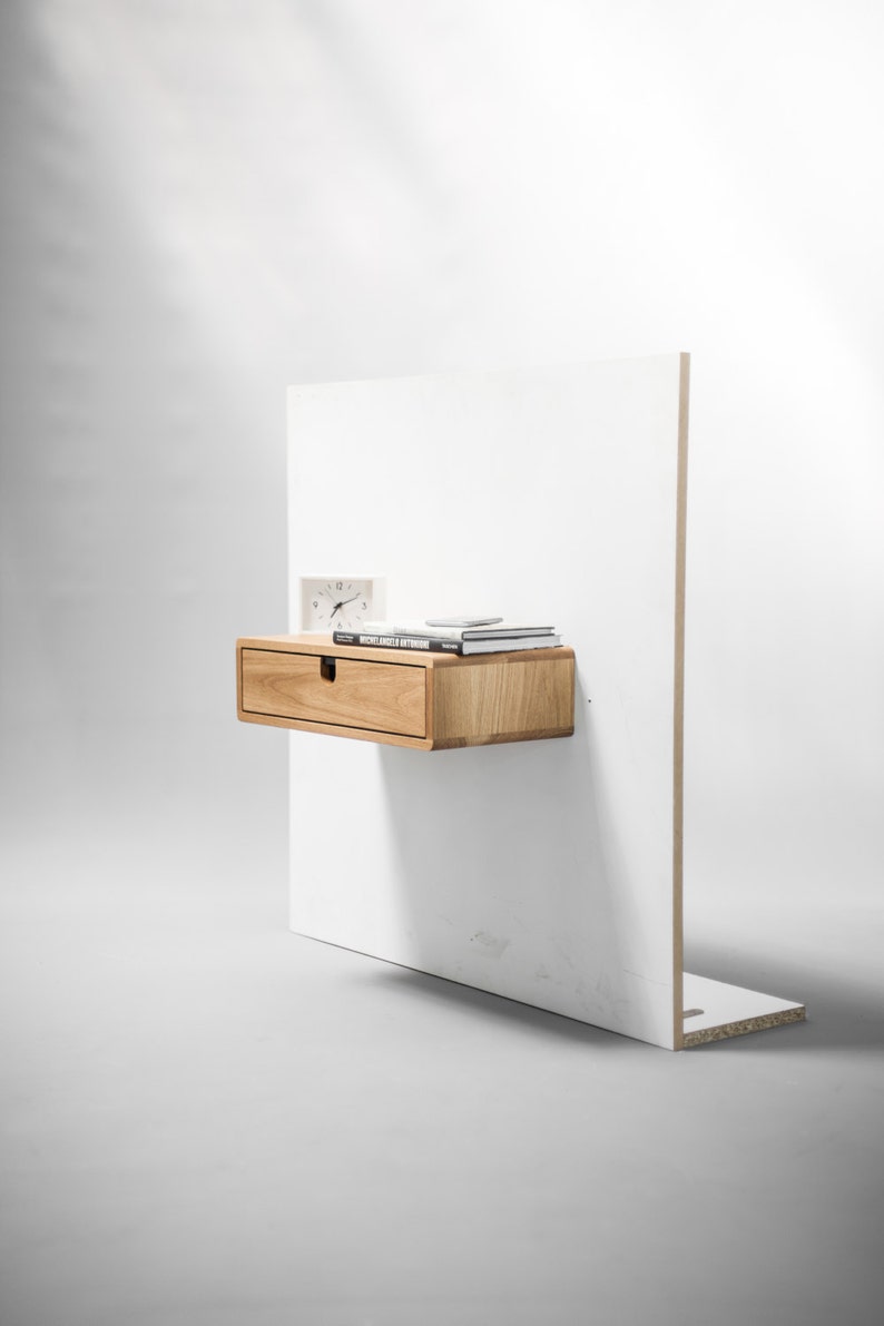 May include: A light wood floating nightstand with a single drawer and a white wall behind it. The nightstand has a clock and books on top.