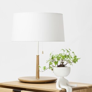 May include: A white table lamp with a white shade sits on a wooden nightstand with two drawers. A white ceramic planter shaped like a person with a green plant inside sits on the nightstand. The text "Manuel Barroso Habitables" is visible on the nightstand.