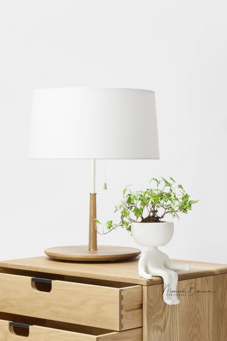 May include: A white table lamp with a white shade sits on a wooden nightstand with two drawers. A white ceramic planter shaped like a person with a green plant inside sits on the nightstand. The text "Manuel Barroso Habitables" is visible on the nightstand.