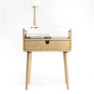 May include: A white and natural wood end table with a single drawer and a shelf. The table has a white lamp with a round white shade on top of it. The table is on a white background.