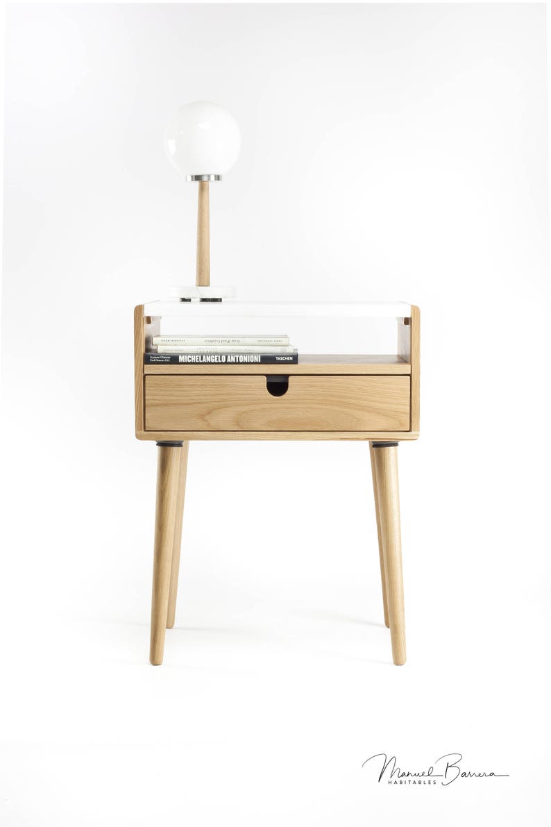 May include: A white and natural wood end table with a single drawer and a shelf. The table has a white lamp with a round white shade on top of it. The table is on a white background.