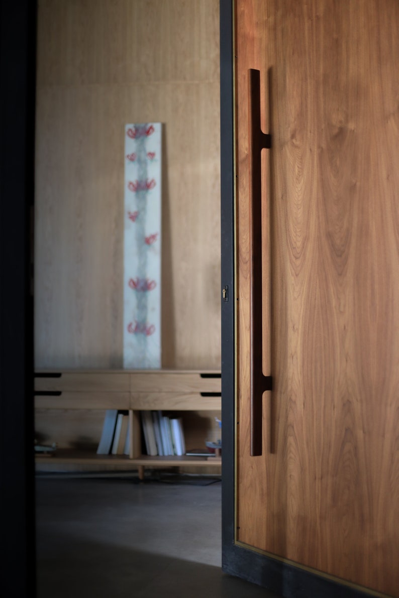 May include: A wooden door with a long, dark brown handle. The door is closed and has a black frame. The door is made of wood with a natural grain pattern.