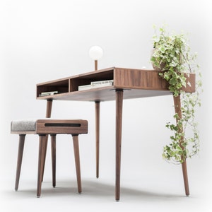 May include: A wooden desk with a light wood finish and a gray upholstered stool. The desk has a shelf and a plant on top. The text "Dressing in Blue Days" is visible on a book on the desk.