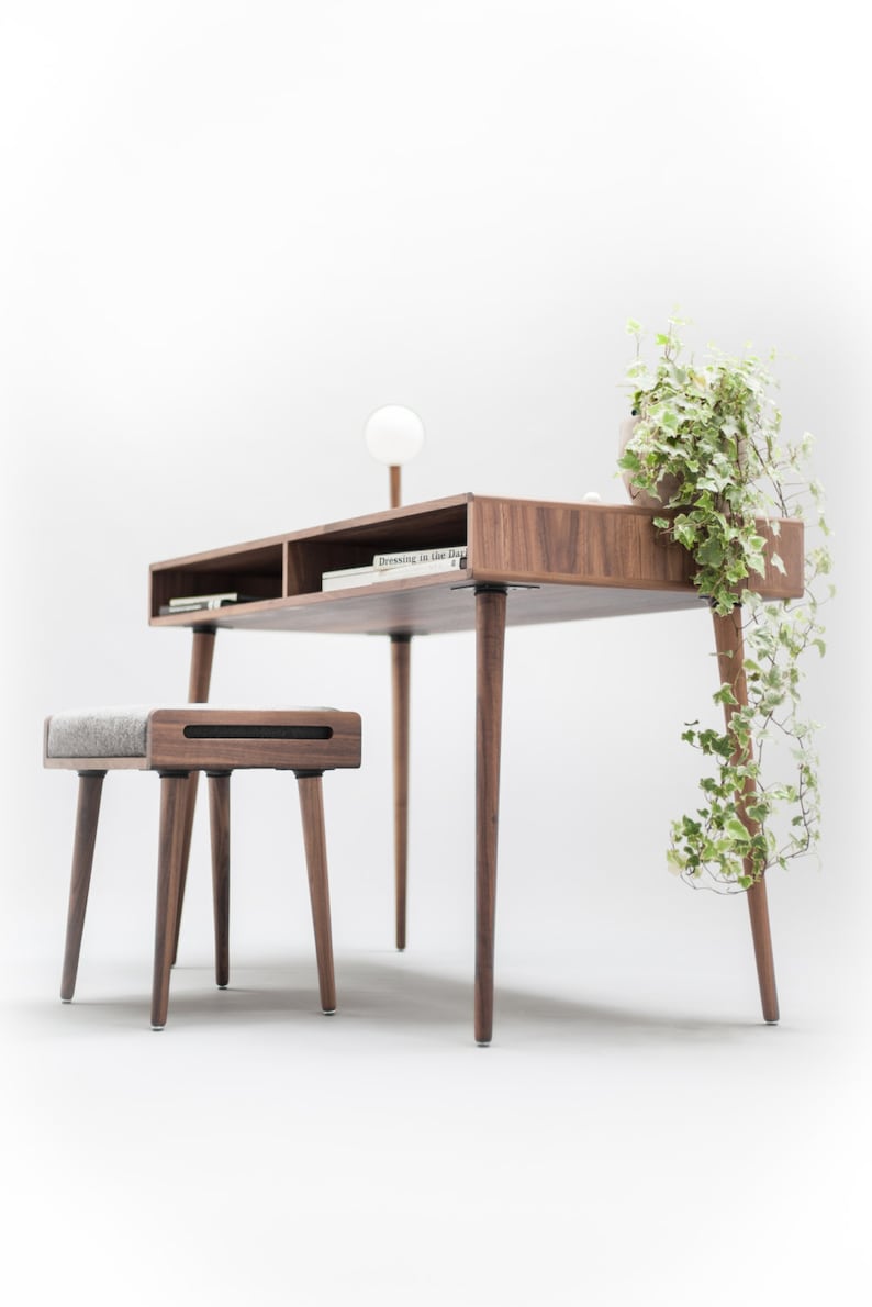 May include: A wooden desk with a light wood finish and a gray upholstered stool. The desk has a shelf and a plant on top. The text "Dressing in Blue Days" is visible on a book on the desk.