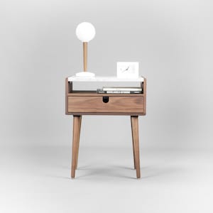 May include: A white marble top nightstand with a single drawer and tapered wooden legs. A white lamp with a round shade and a white clock are on the top shelf. A book titled "Michelangelo Antonioni" is on the lower shelf.
