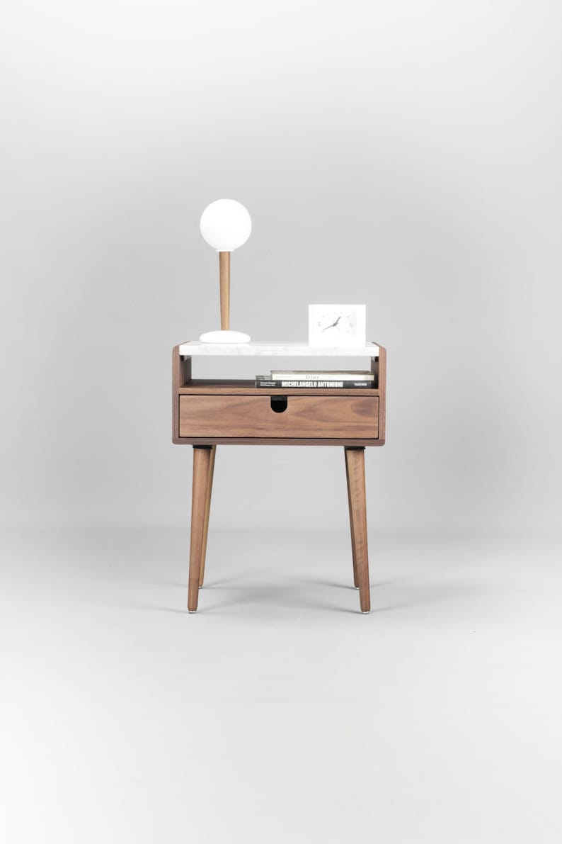 May include: A white marble top nightstand with a single drawer and tapered wooden legs. A white lamp with a round shade and a white clock are on the top shelf. A book titled "Michelangelo Antonioni" is on the lower shelf.
