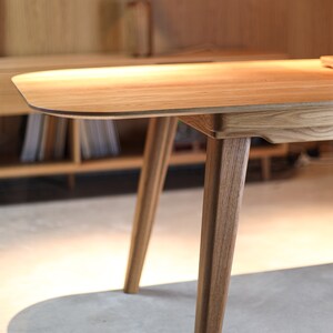 May include: A close-up of a wooden table with a light brown finish. The table has a rounded edge and four legs. The legs are made of solid wood and have a natural grain.