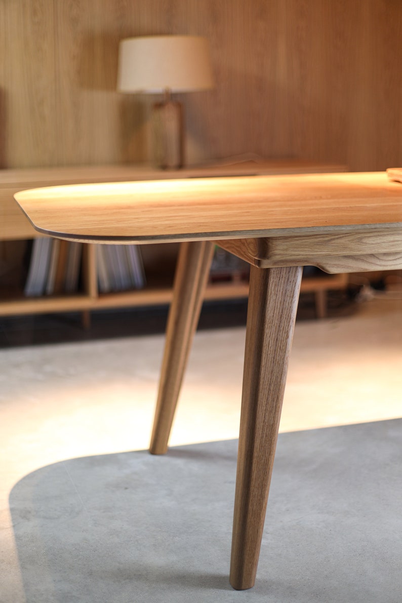 May include: A close-up of a wooden table with a light brown finish. The table has a rounded edge and four legs. The legs are made of solid wood and have a natural grain.