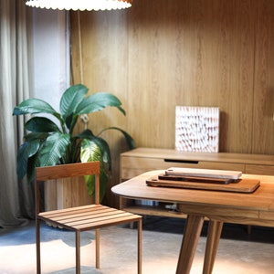 May include: A wooden table with a light wood finish and a rounded edge. The table has three legs and is set with two wooden cutting boards. A wooden chair with a slatted seat and back is positioned to the left of the table.