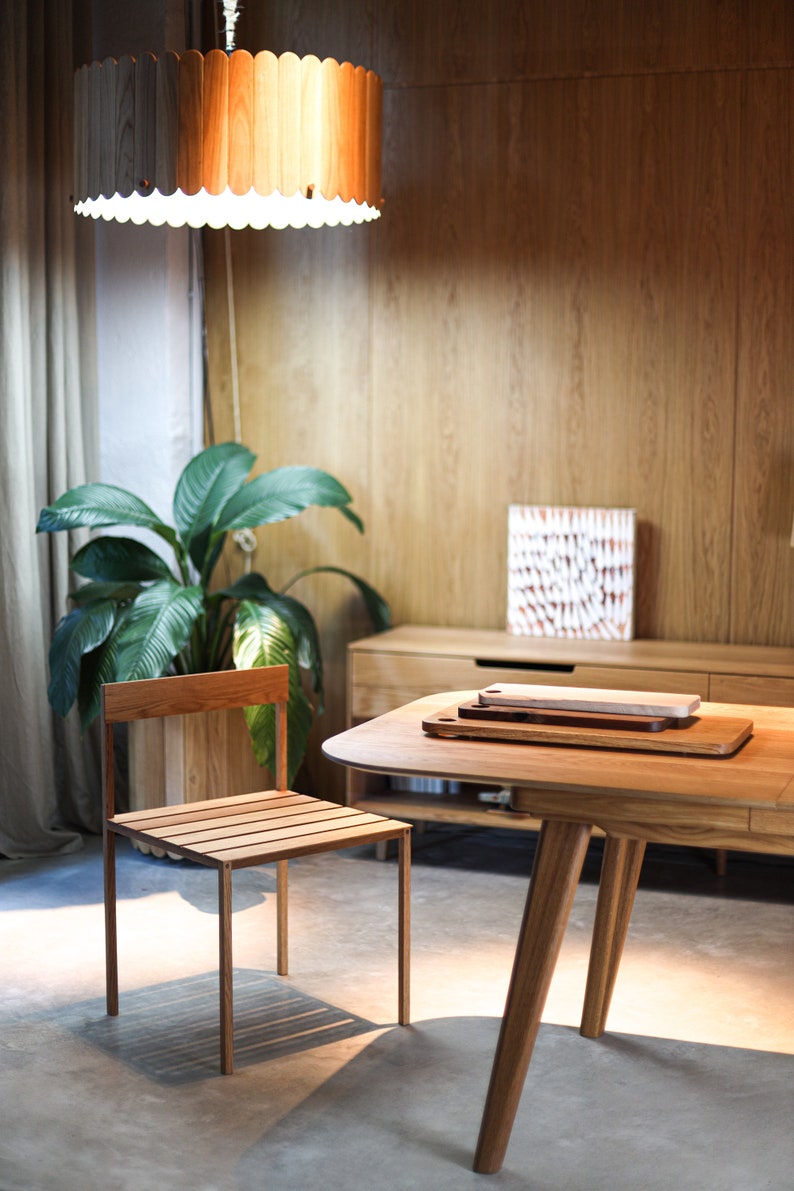 May include: A wooden table with a light wood finish and a rounded edge. The table has three legs and is set with two wooden cutting boards. A wooden chair with a slatted seat and back is positioned to the left of the table.