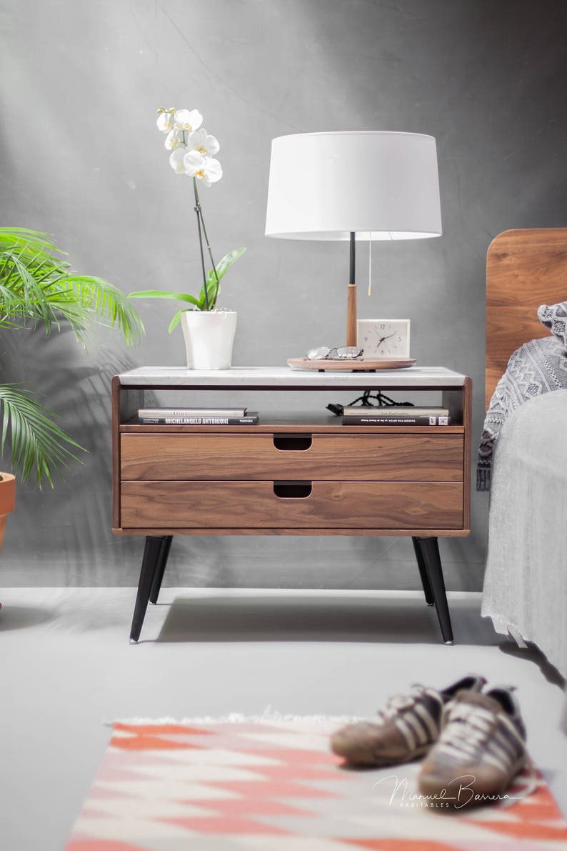May include: A wooden nightstand with two drawers and black legs. The nightstand has a white marble top and a shelf underneath the top. A white lamp with a white shade is on the nightstand. A potted white orchid is on the left side of the nightstand. A clock is on the nightstand.