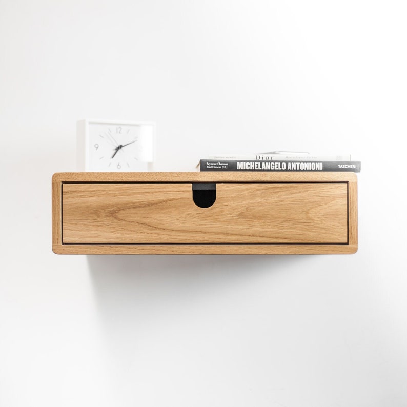 May include: A light wood floating shelf with a single drawer and a black pull. A white clock and a book titled "Michelangelo Antonioni" are on top of the shelf.