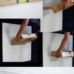 May include: A light wood floating shelf with a white drawer is being installed on a white wall. A level is used to ensure the shelf is straight. The shelf is being held up by a person's hands.