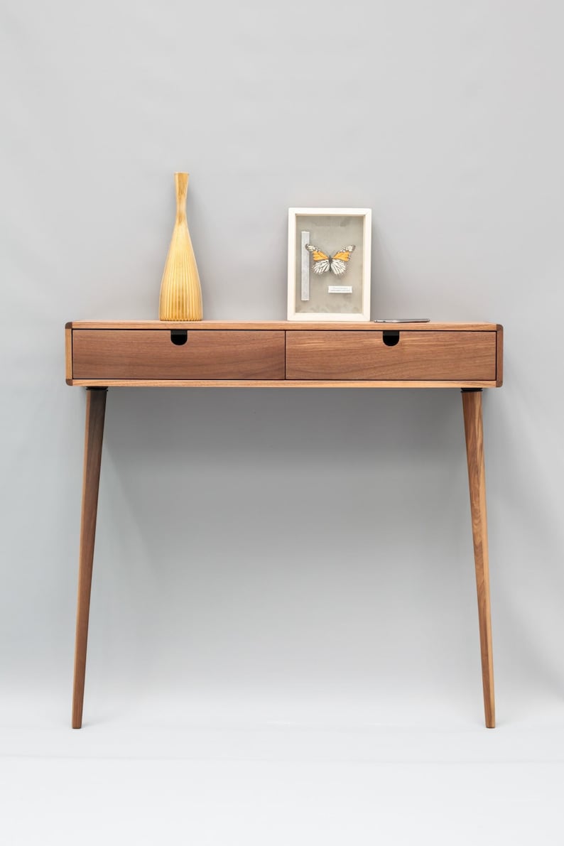 May include: A wooden console table with two drawers and tapered legs. A vase and a framed butterfly specimen are on top of the table.