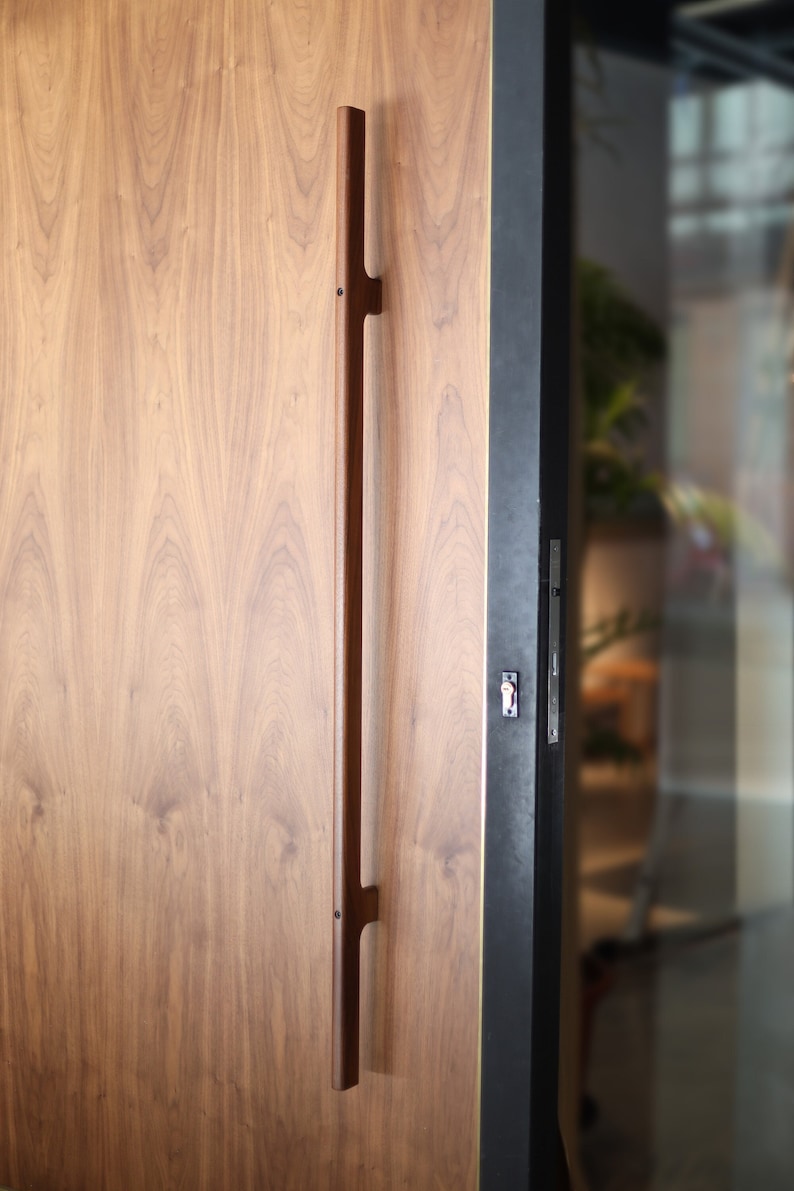 May include: A wooden door with a long, dark brown handle. The door is made of wood with a natural grain pattern. The handle is attached to the door with two small screws.