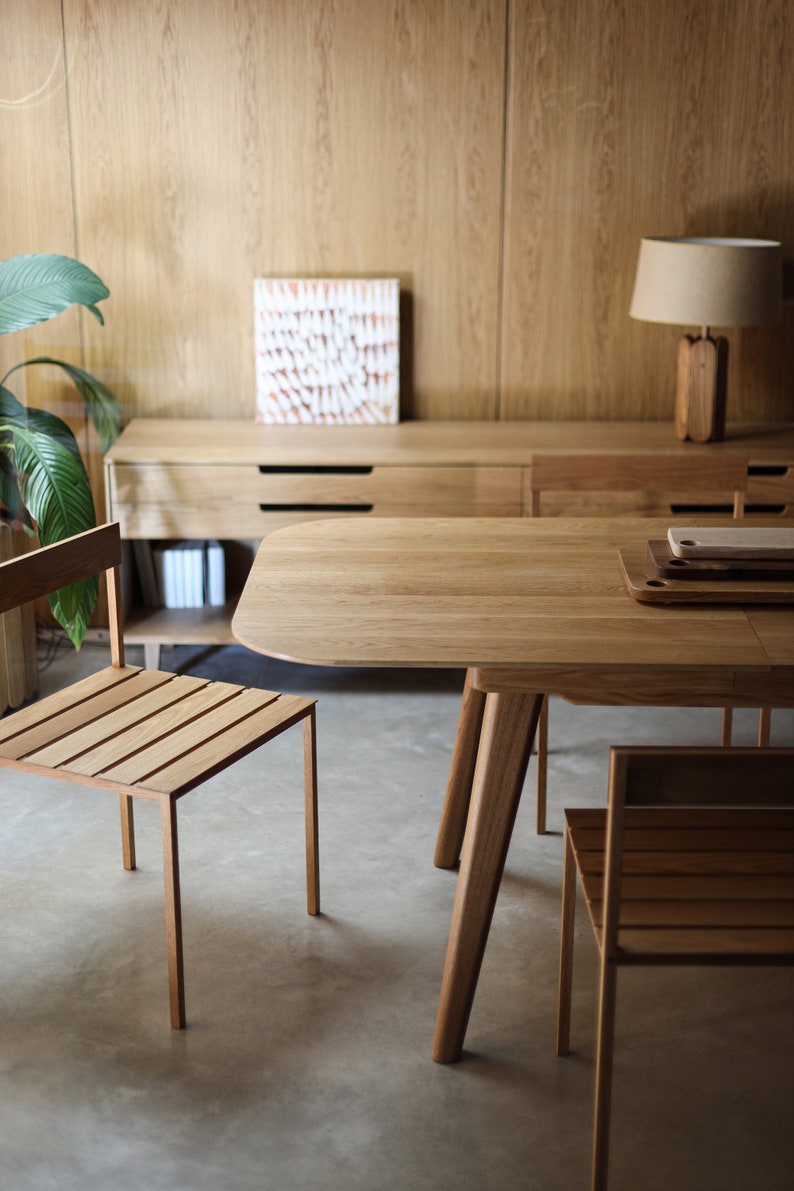 May include: A wooden dining table with a rounded edge and a light wood finish. Two matching wooden chairs with slatted seats are positioned on either side of the table. The table and chairs are in a room with wood paneling on the walls.