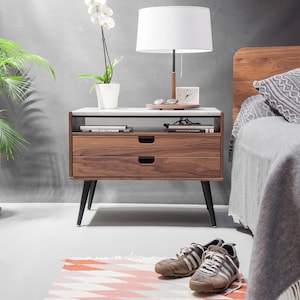 May include: A wooden nightstand with two drawers and black legs. The nightstand has a white marble top and is decorated with a lamp, a clock, and a potted orchid.
