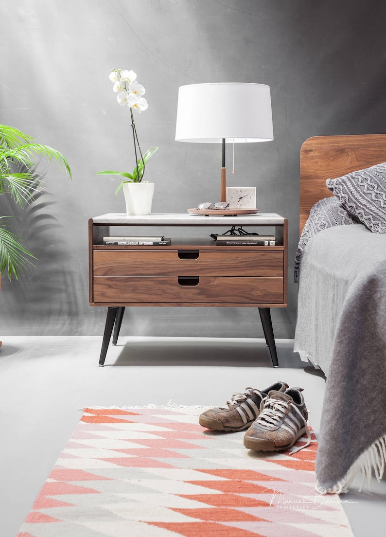 May include: A wooden nightstand with two drawers and black legs. The nightstand has a white marble top and is decorated with a lamp, a clock, and a potted orchid.