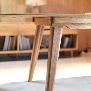 May include: A close-up of a wooden table with a light wood finish. The table has a round top and four legs. The legs are tapered and have a slightly curved shape. The table is sitting on a light-colored floor.