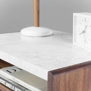 May include: A wooden nightstand with a white marble top. The nightstand has a drawer and a shelf. There is a lamp, a clock, and a stack of books on the nightstand.