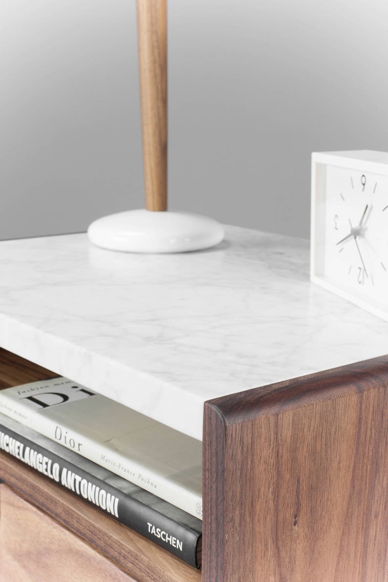 May include: A wooden nightstand with a white marble top. The nightstand has a drawer and a shelf. There is a lamp, a clock, and a stack of books on the nightstand.