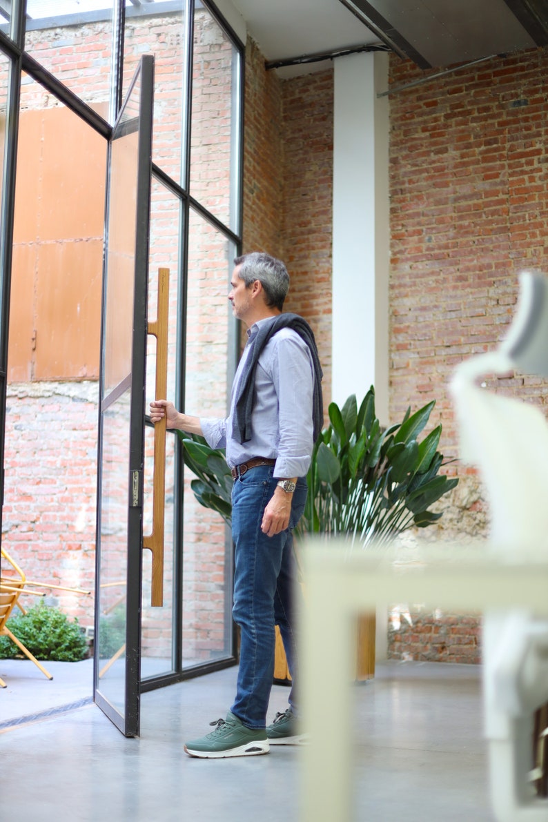 May include: A man in a blue shirt and jeans stands in a room with a brick wall and large windows. He is holding a wooden door handle and looking out the window.