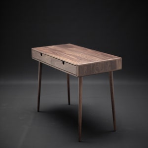 May include: A dark brown wooden desk with two drawers and tapered legs. The desk is on a black background.