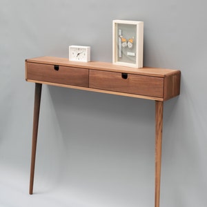 May include: A wooden console table with two drawers and tapered legs. The table is made of walnut wood and has a natural finish. A small white clock and a framed picture of a butterfly are on top of the table.