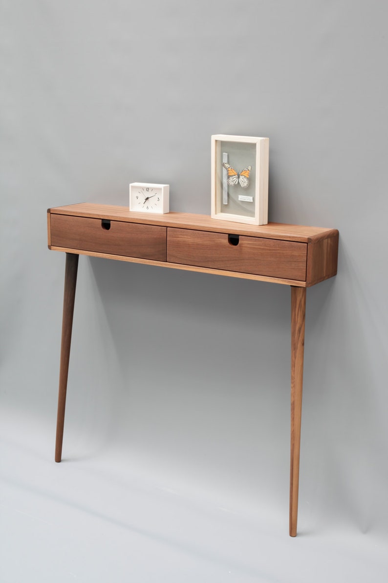 May include: A wooden console table with two drawers and tapered legs. The table is made of walnut wood and has a natural finish. A small white clock and a framed picture of a butterfly are on top of the table.